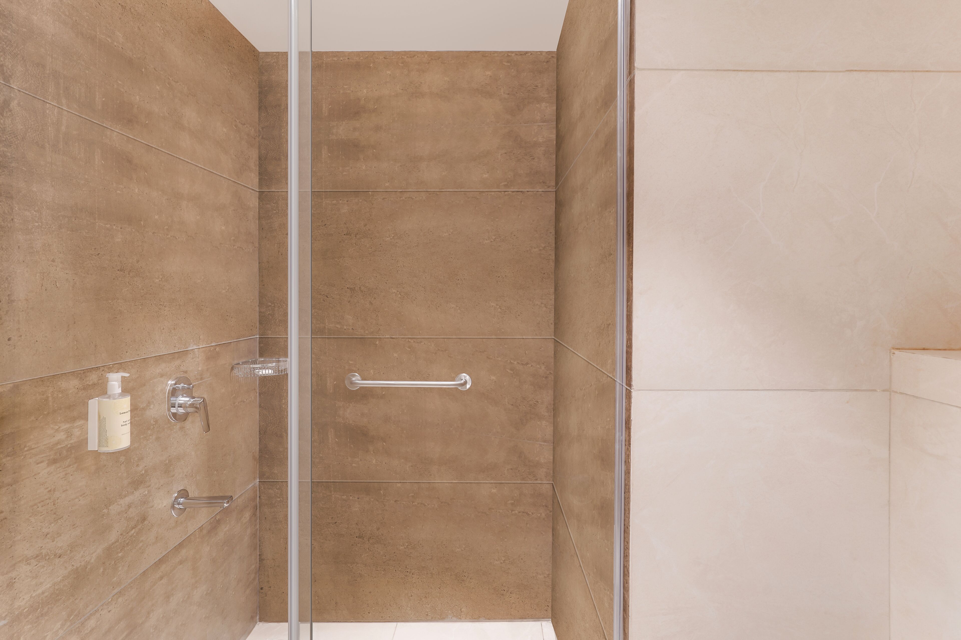 A modern bathroom with a glass shower enclosure, beige tiles, and a handrail.