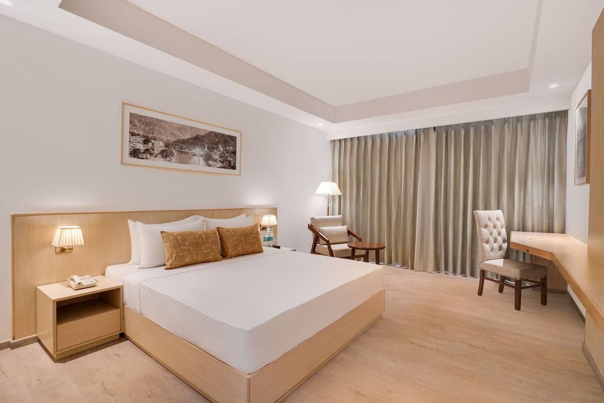 A hotel room with a large bed, bedside tables with lamps, a desk, a chair, and a large window with curtains.