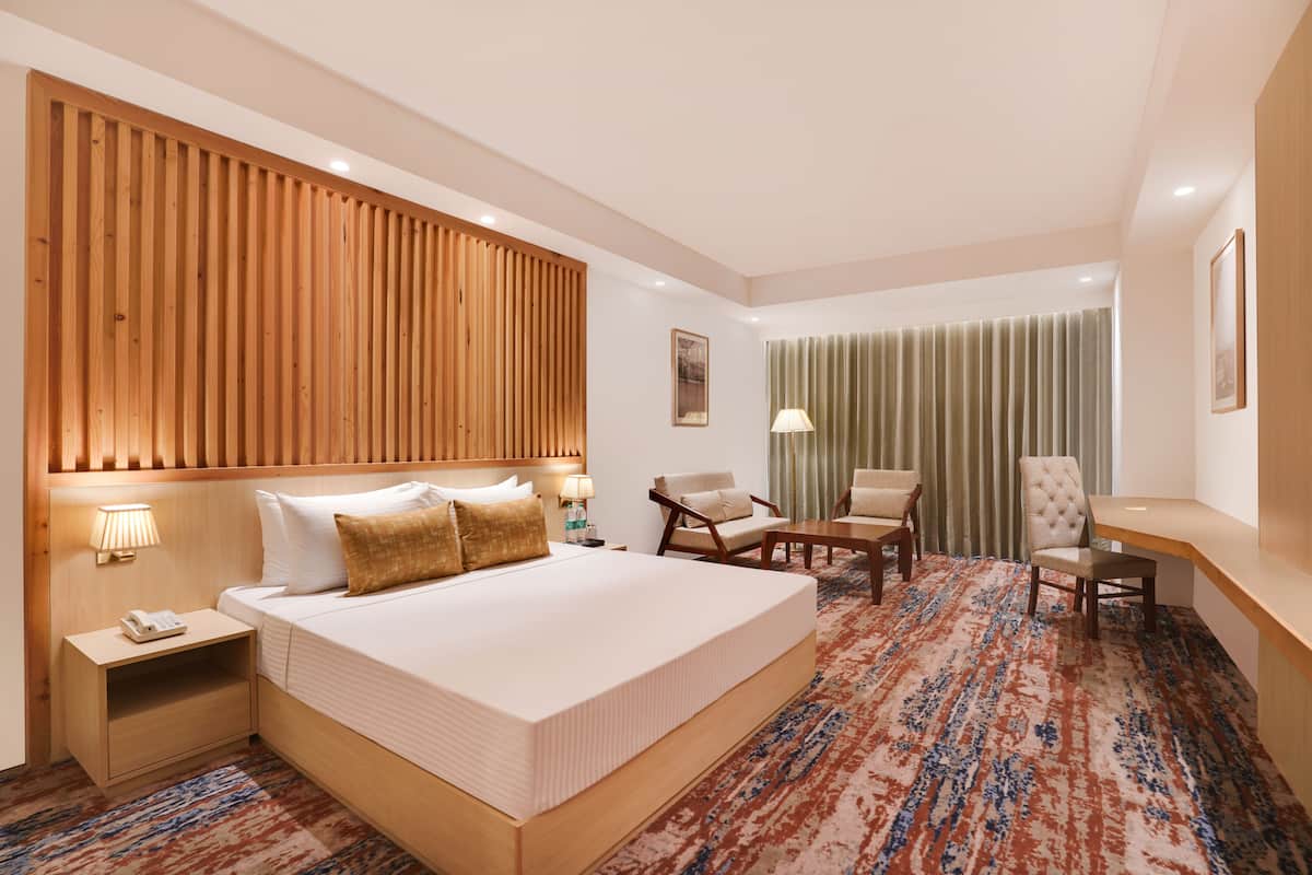 A hotel room with a large bed, a wooden headboard, a desk, two chairs, a nightstand with a phone, and a patterned carpet.