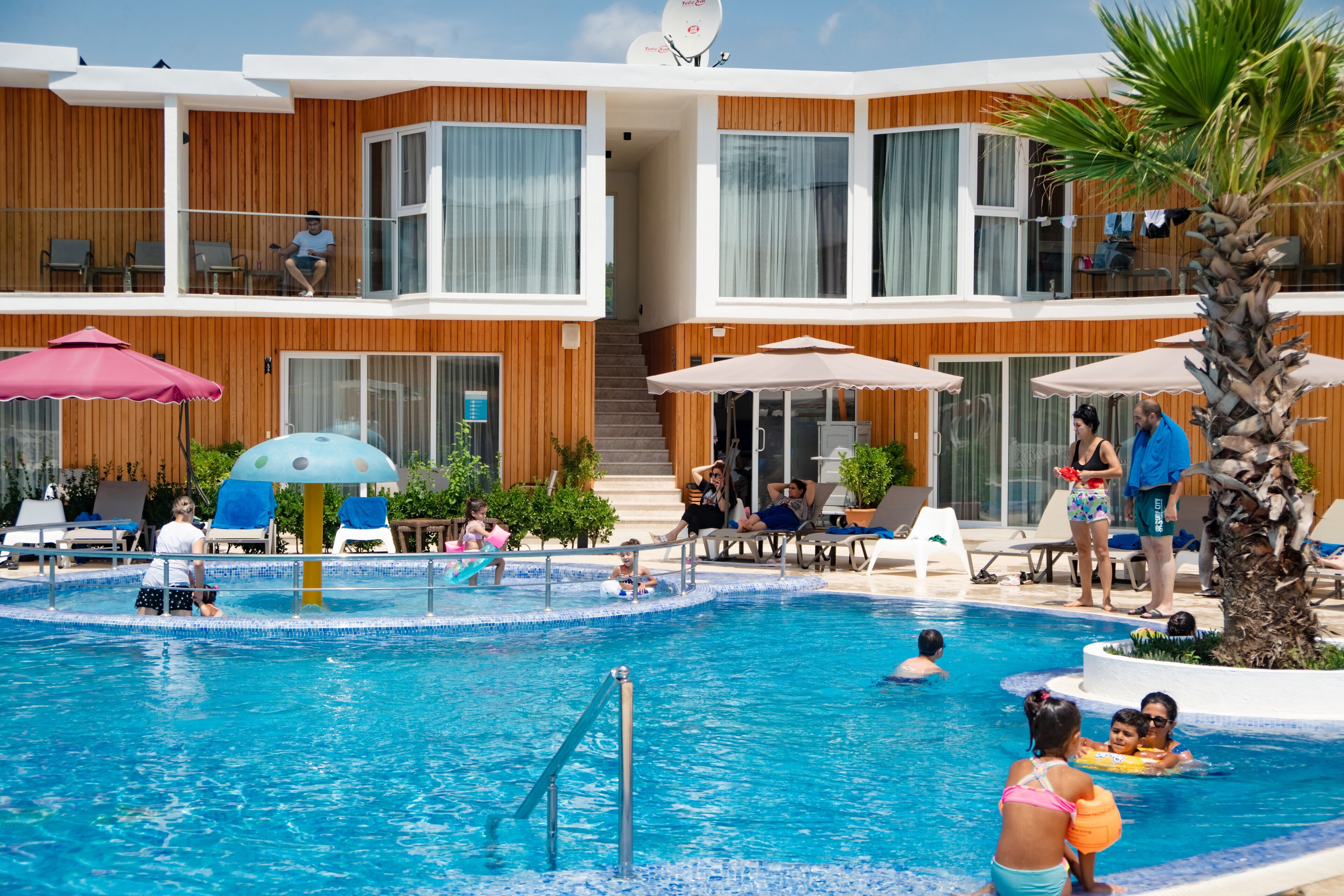 A swimming pool area with lounge chairs, umbrellas, and a multi-level building with wooden paneling and large windows.