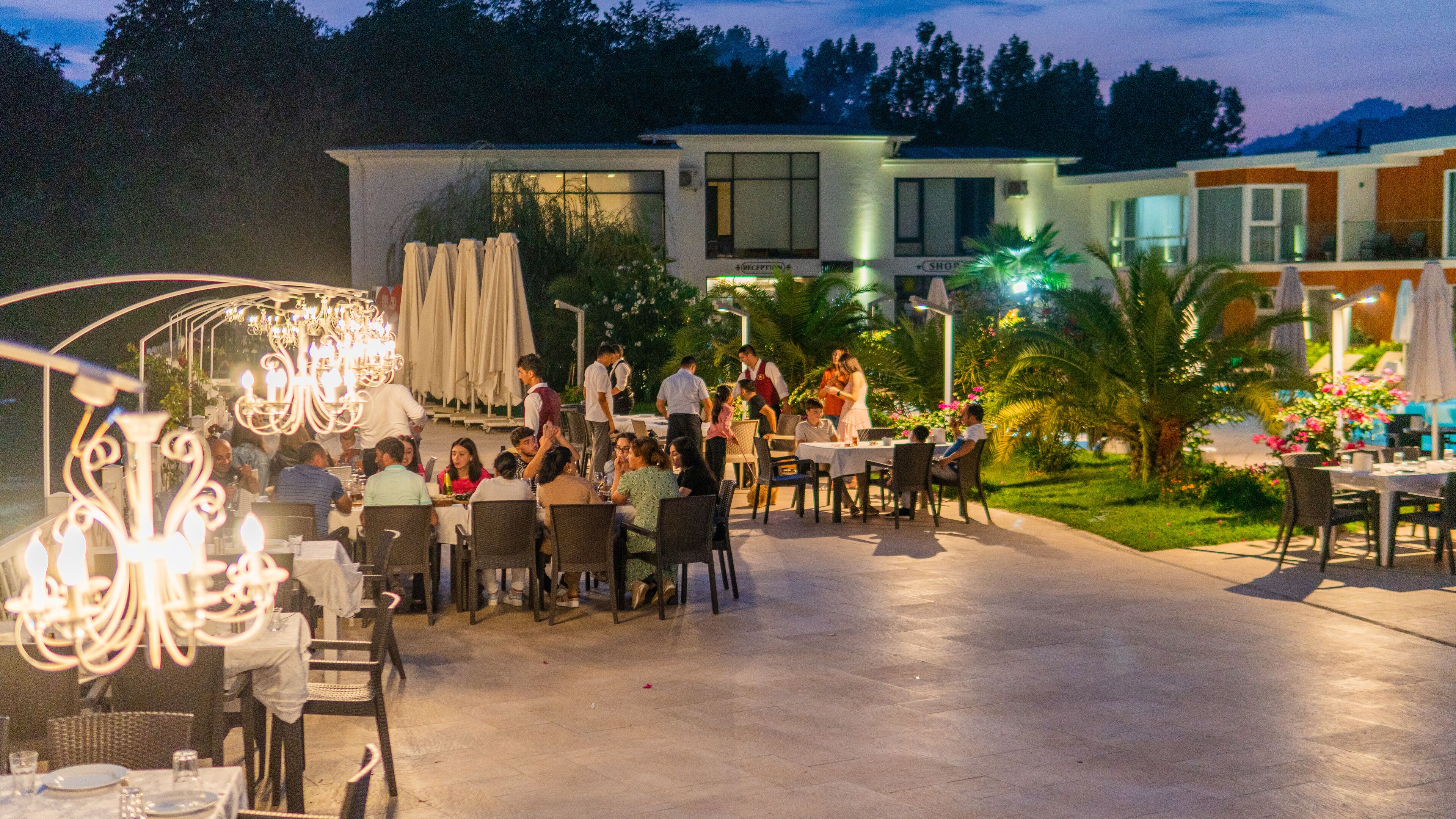 An outdoor dining area with tables and chairs, a reception area, and a building with a shop sign in the background.