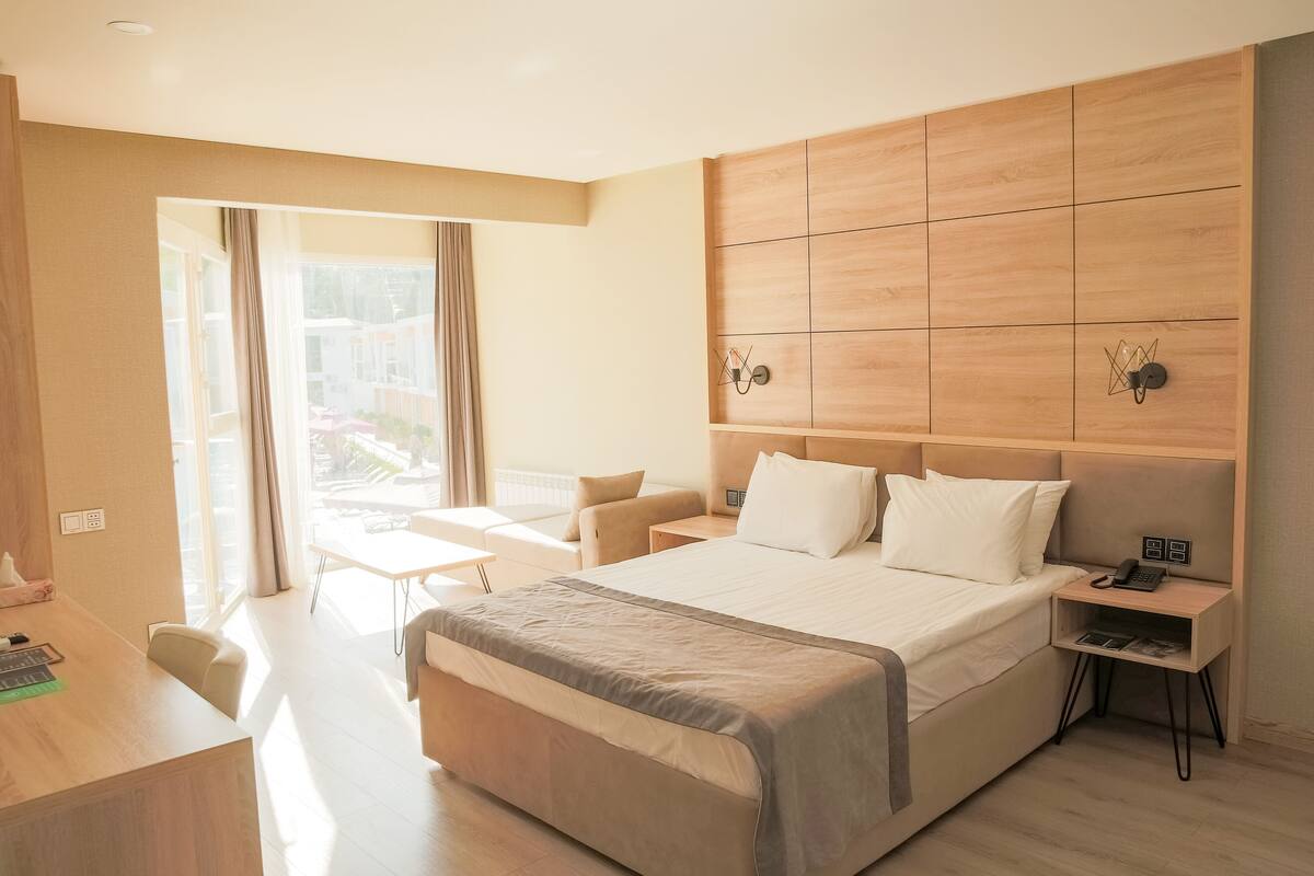 A modern hotel room with a large bed, a desk, and a chair. There is a window with curtains, a bedside table with a lamp, and a wall-mounted phone.