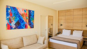 A hotel room with a beige sofa, a bed with a wooden headboard, a colorful abstract painting on the wall, and a wooden floor.