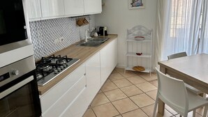 A modern kitchen with white cabinets, a gas stove, a microwave, and a sink. There's a white shelf with a bowl and a red bowl on it. A dining table with chairs is visible.