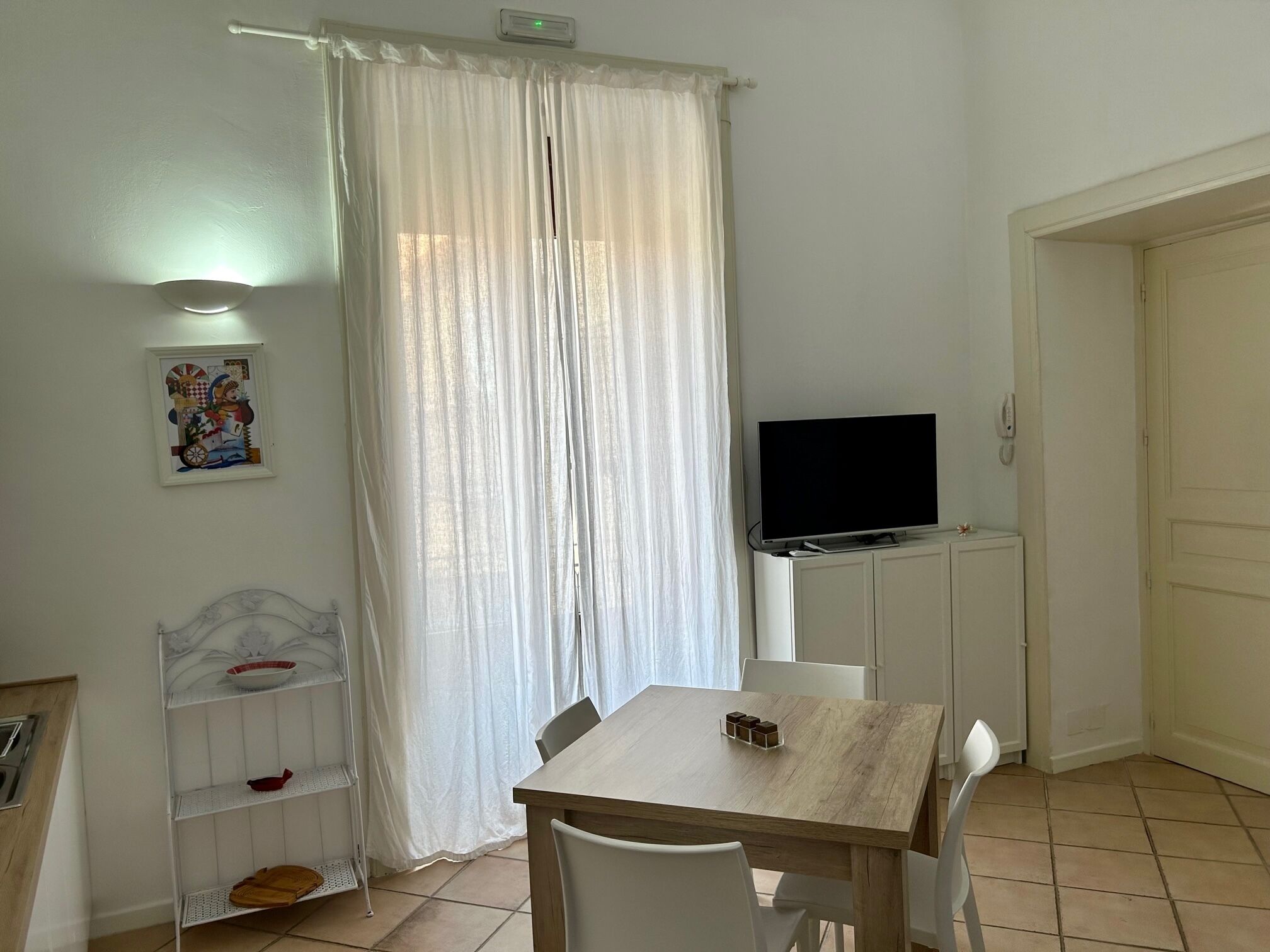 A room with a wooden table, white chairs, a TV on a stand, a shelf with items, a door, and a curtain.