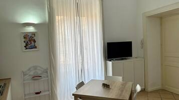 A room with a wooden table, white chairs, a TV on a stand, a shelf with items, a door, and a curtain.