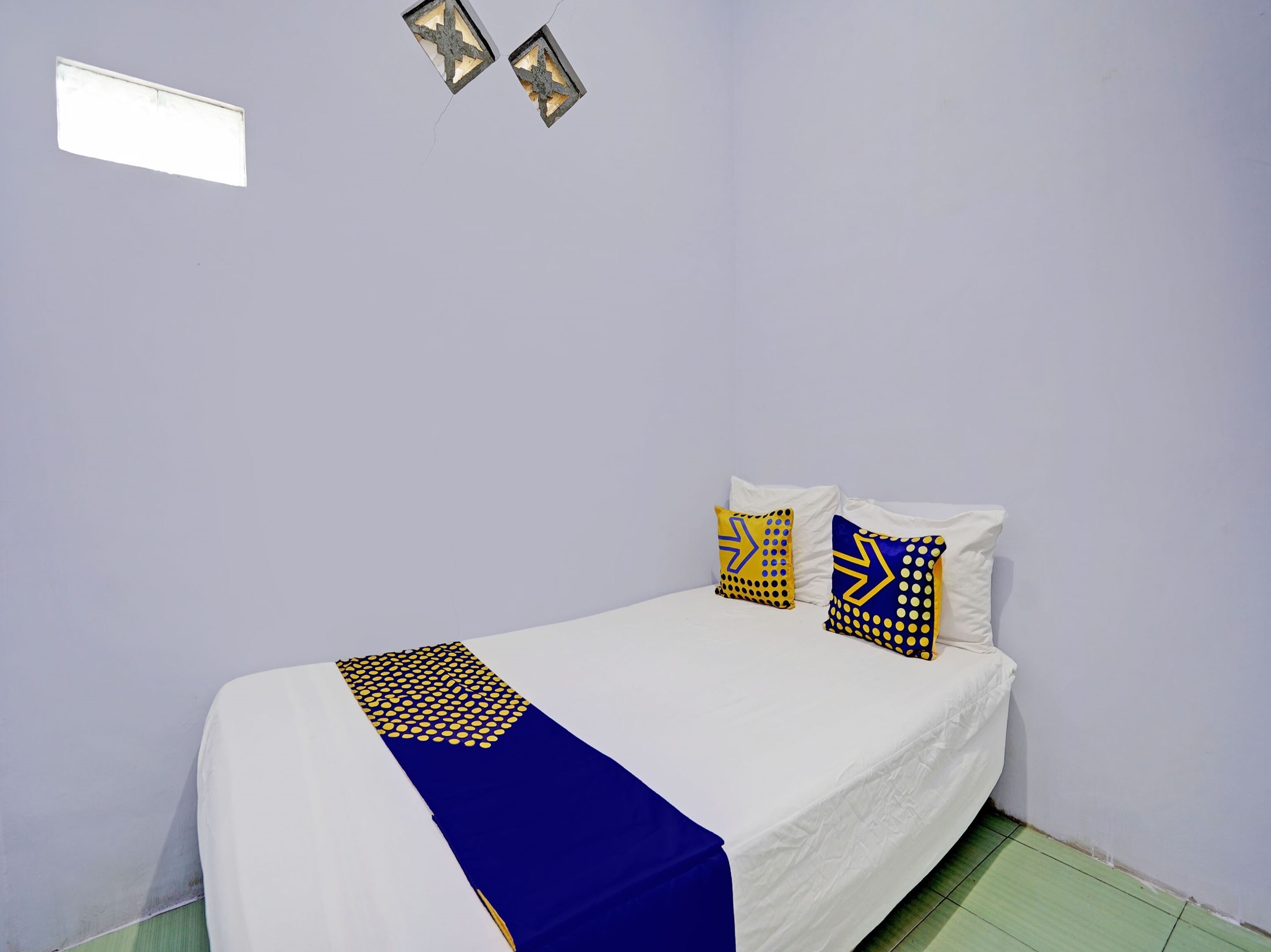 Uma cama de solteiro com colchão branco e colcha com padrão azul e amarelo. Há duas almofadas com estampas em azul e amarelo. O quarto tem paredes lilases claras e uma pequena janela com luminária.