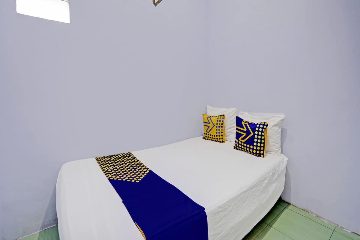 A single bed with a white mattress and blue and yellow patterned bedspread. There are two pillows with yellow and blue patterns. The room has light purple walls and a small window with a light fixture.