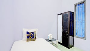A bedroom with a bed, a bedside table with a fan, a wardrobe, and a window with blue curtains. - OYO Life 92937 Kost Putri Sarkara Syariah (Rogojampi)