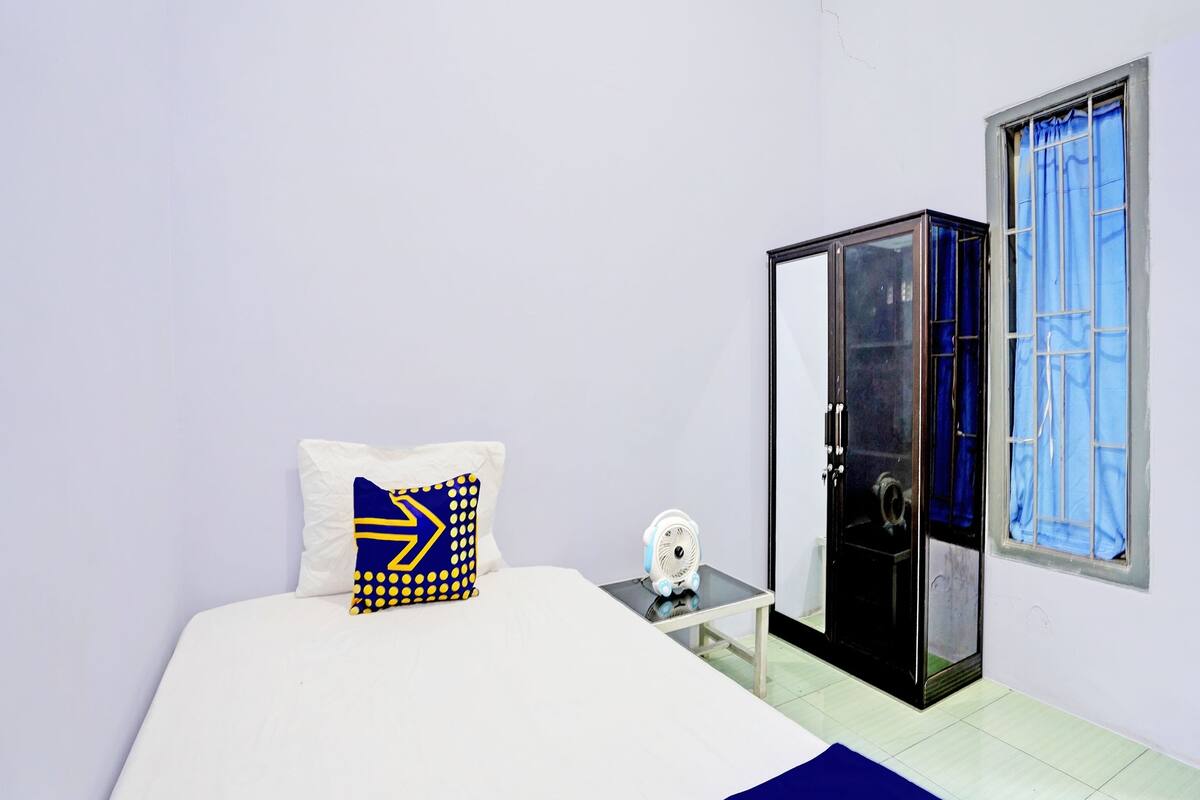 A bedroom with a bed, a bedside table with a fan, a wardrobe, and a window with blue curtains.