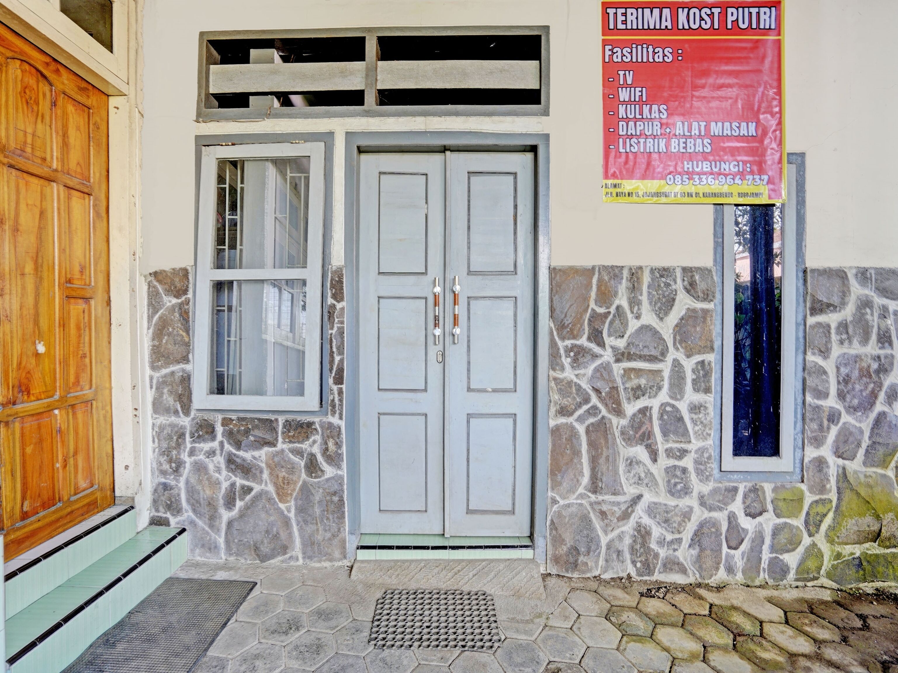 Entrada de um edifício com porta de madeira, parede de pedra e placa listando comodidades como TV, WiFi e cozinha.