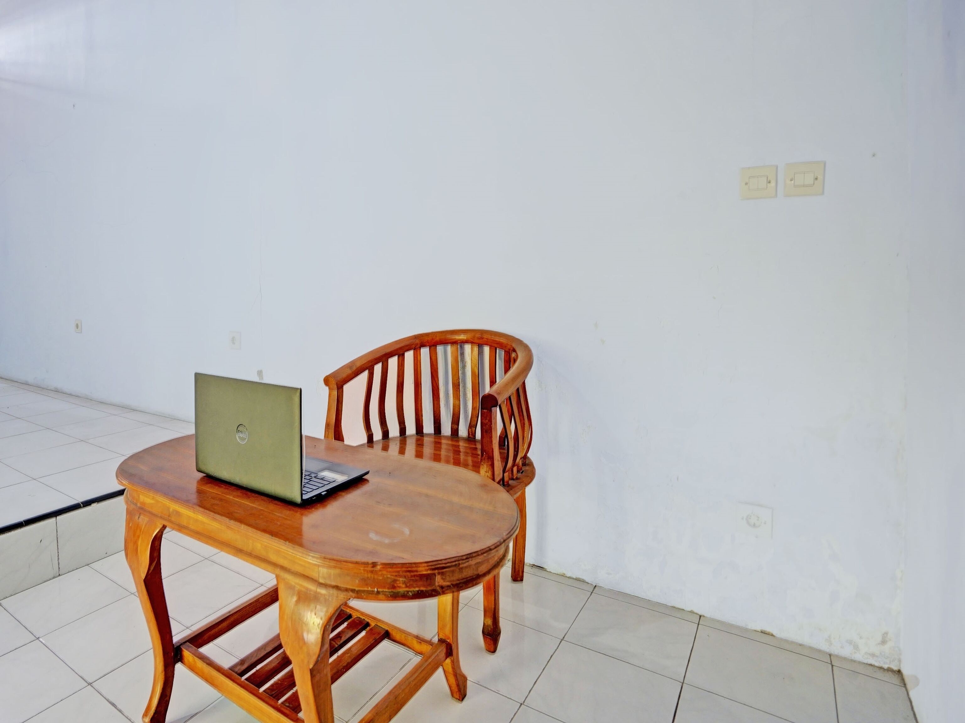 Een houten tafel met een laptop en een stoel in een kamer met witte wanden en betegelde vloer.