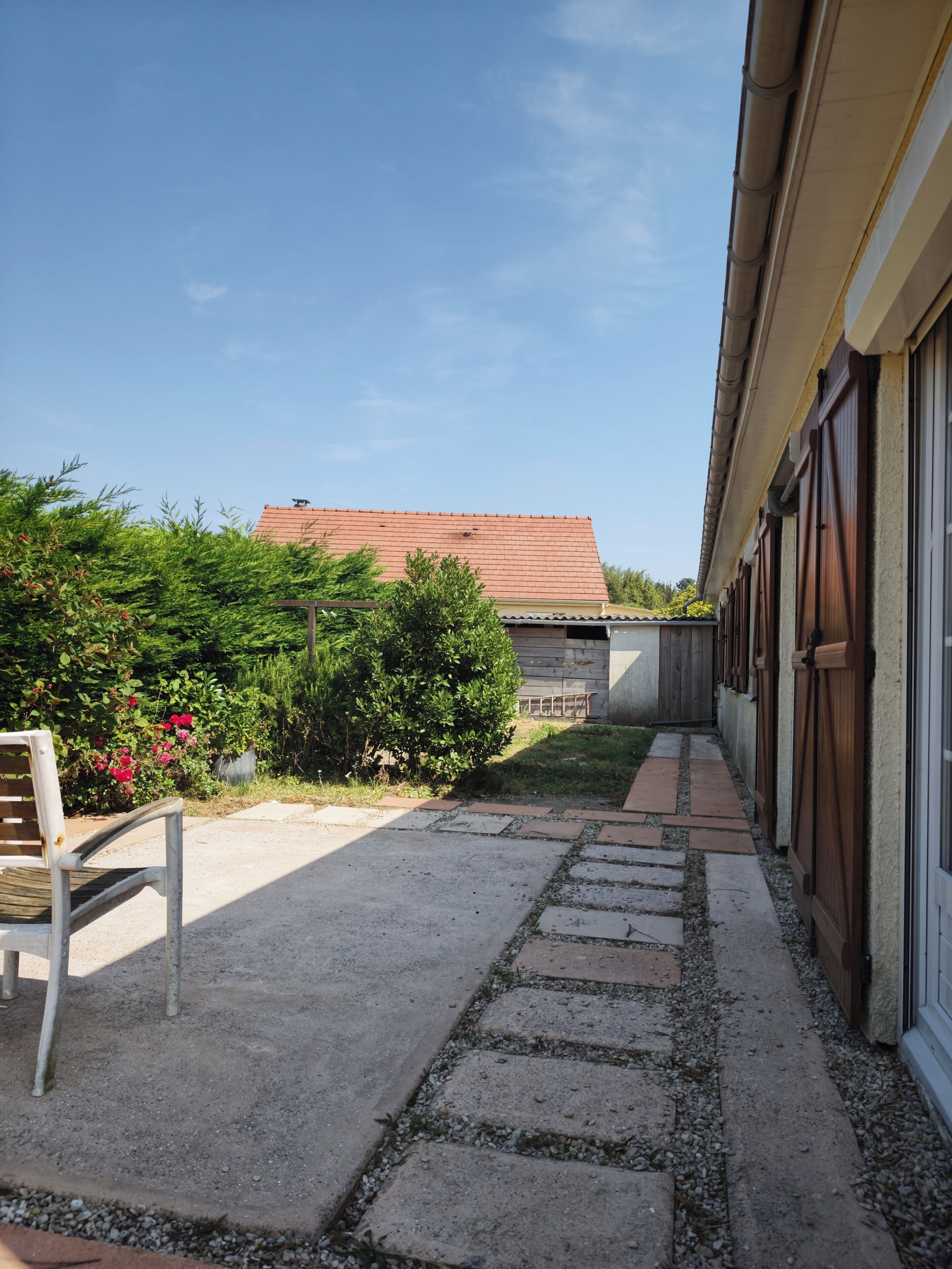 A paved pathway leading to a building with a red roof, surrounded by greenery and a bench.