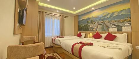 A hotel room with two beds, a large mural of a village, a wooden chair, and a desk with a lamp.