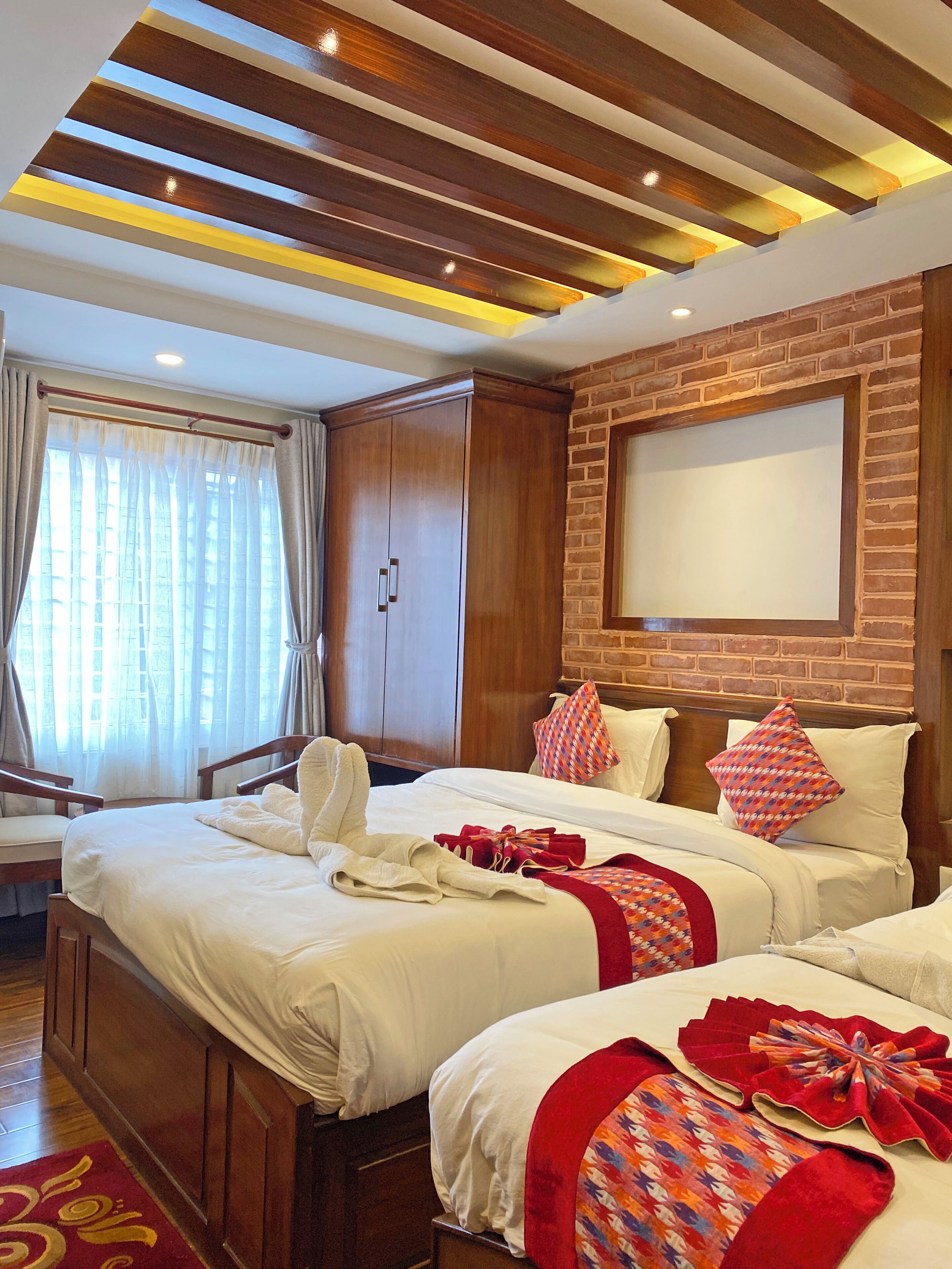 Habitación de hotel con techo de madera, pared de ladrillo, una cama grande con ropa blanca, dos mesitas de noche y una ventana con cortinas.