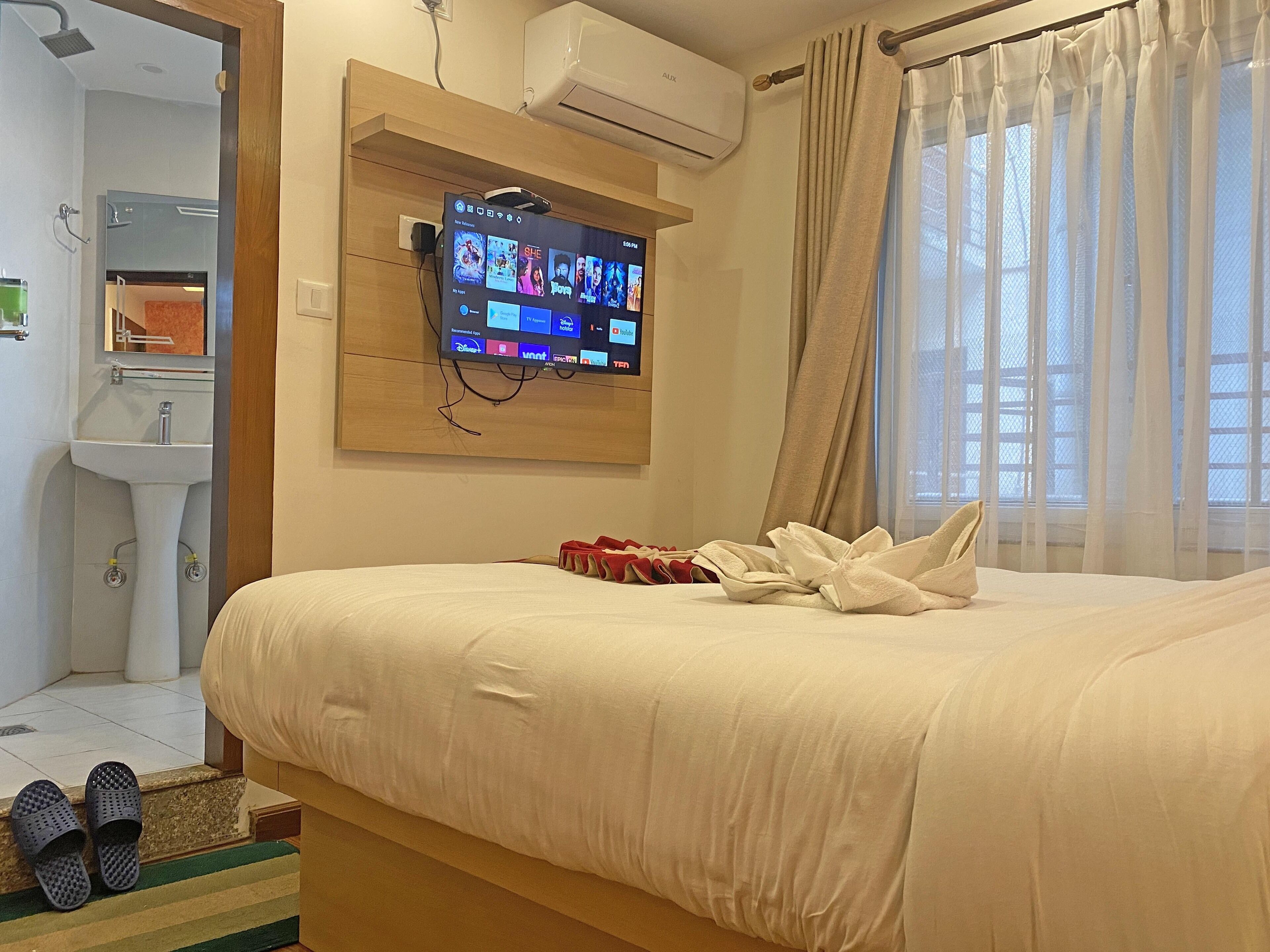 Camera d'albergo con un letto, una TV a muro che mostra diversi canali, un bagno con un lavandino e una finestra con tende.