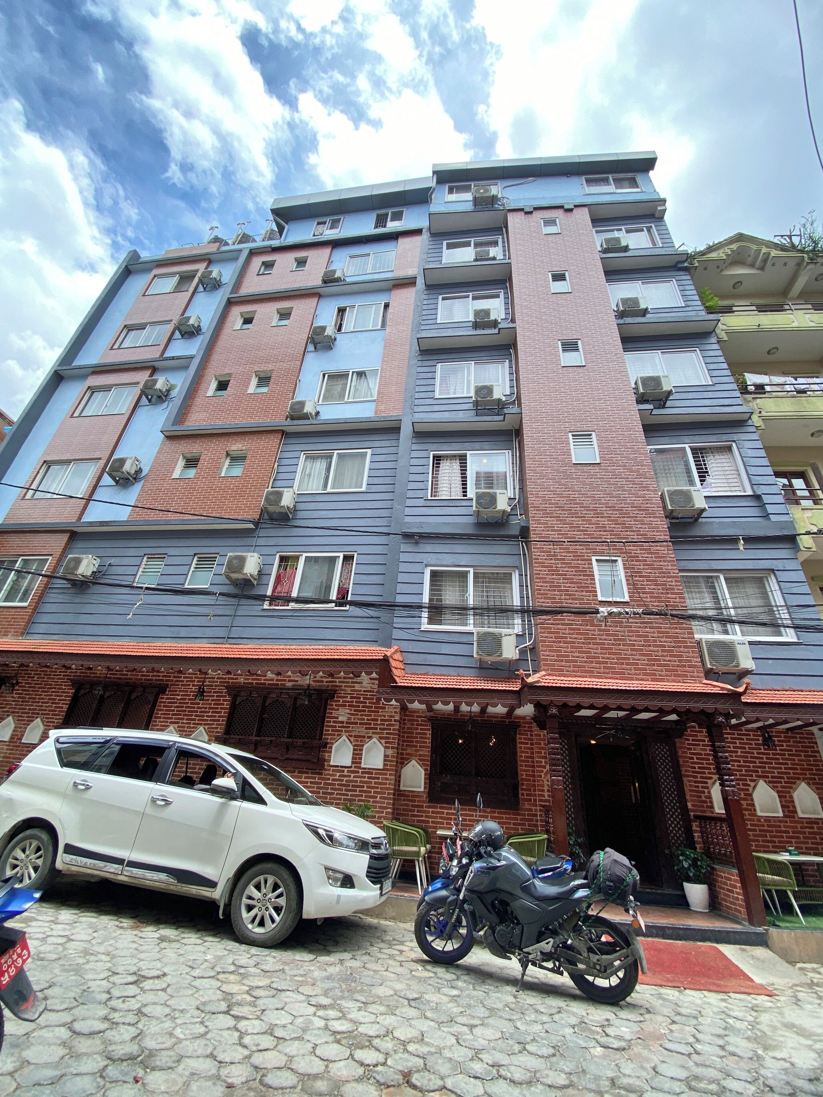 A multi-story residential building with air conditioning units, a car, and a motorcycle parked in front.