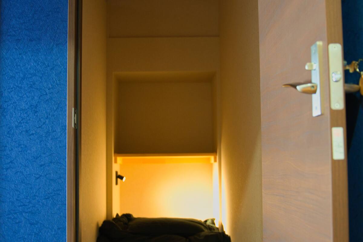 A narrow hallway with wooden flooring leading to a room with a bed and a light source.