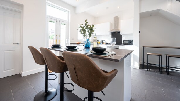A modern kitchen with a central island, two bar stools, and a vase with green leaves. - Durham View by Tŷ SA (Newport)