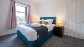 A neatly made bed with a blue headboard, a bedside table with a lamp, and a window with curtains. - Durham View by Tŷ SA (Newport)