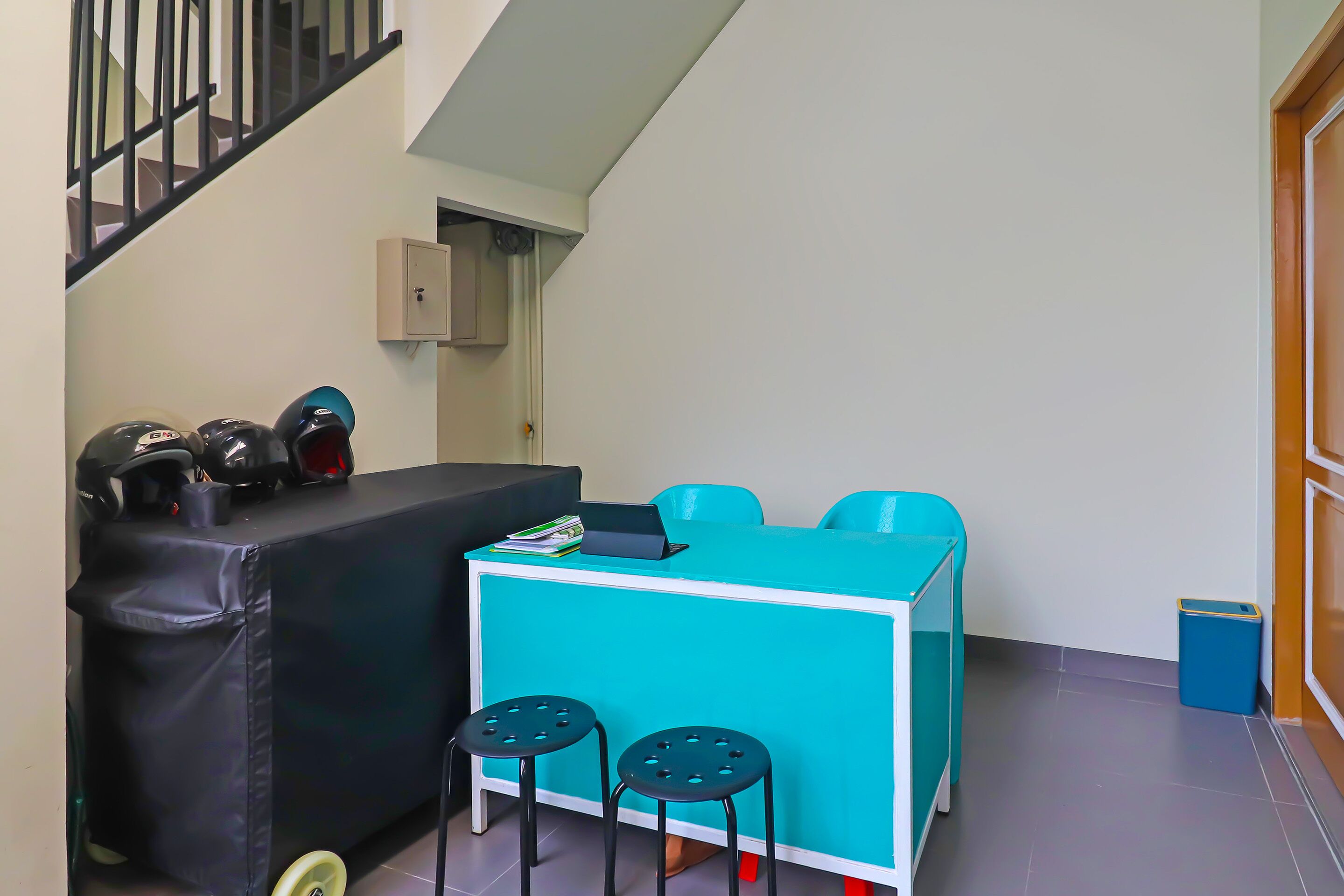 Ein kleines Zimmer mit einem türkisblauen Schreibtisch, zwei schwarzen Hocker, einer schwarzen Couch und einer Treppe.
