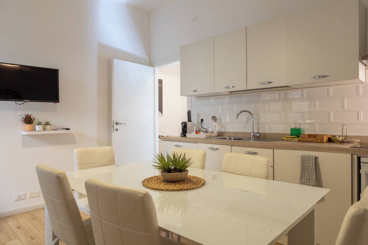 Una cocina moderna con armarios blancos, un televisor de pantalla plana montado sobre la encimera y un comedor con mesa y sillas blancas.