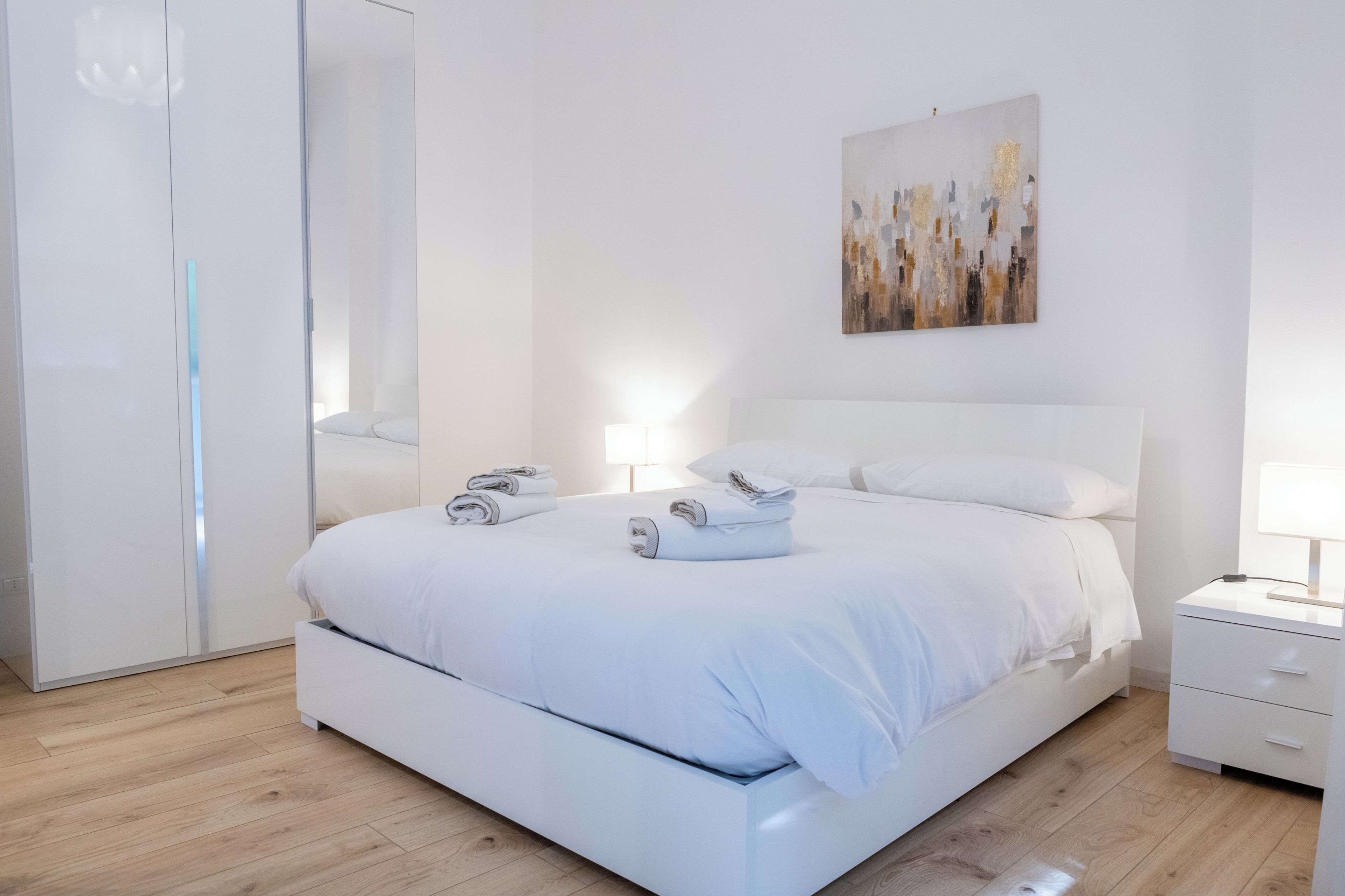 A modern bedroom with a white bed, bedside tables, a nightstand, and a wall-mounted abstract painting.