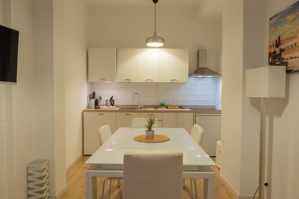Una cocina moderna con armarios blancos, una mesa de comedor con sillas, una lámpara colgante y un cuadro en la pared.