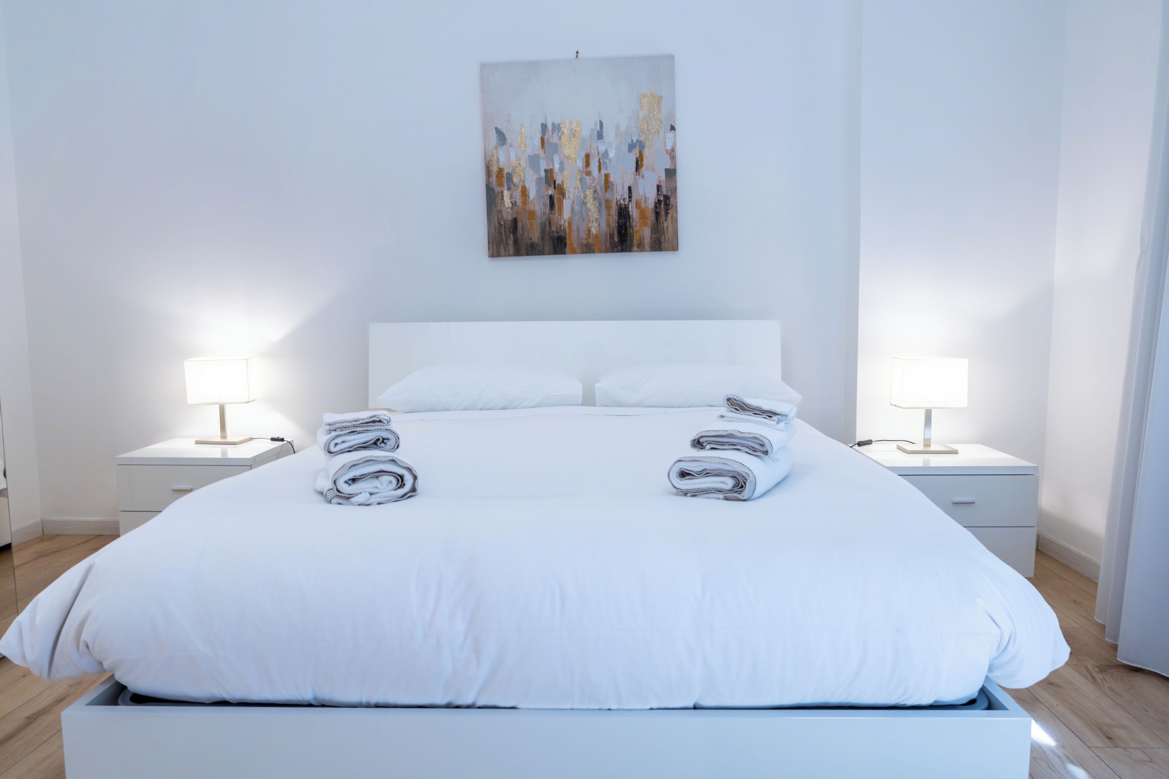 Ein ordentlich bezogenes Bett mit weißen Leinen und Handtüchern, zwei Nachttische mit Lampen sowie ein Bild an der Wand.
