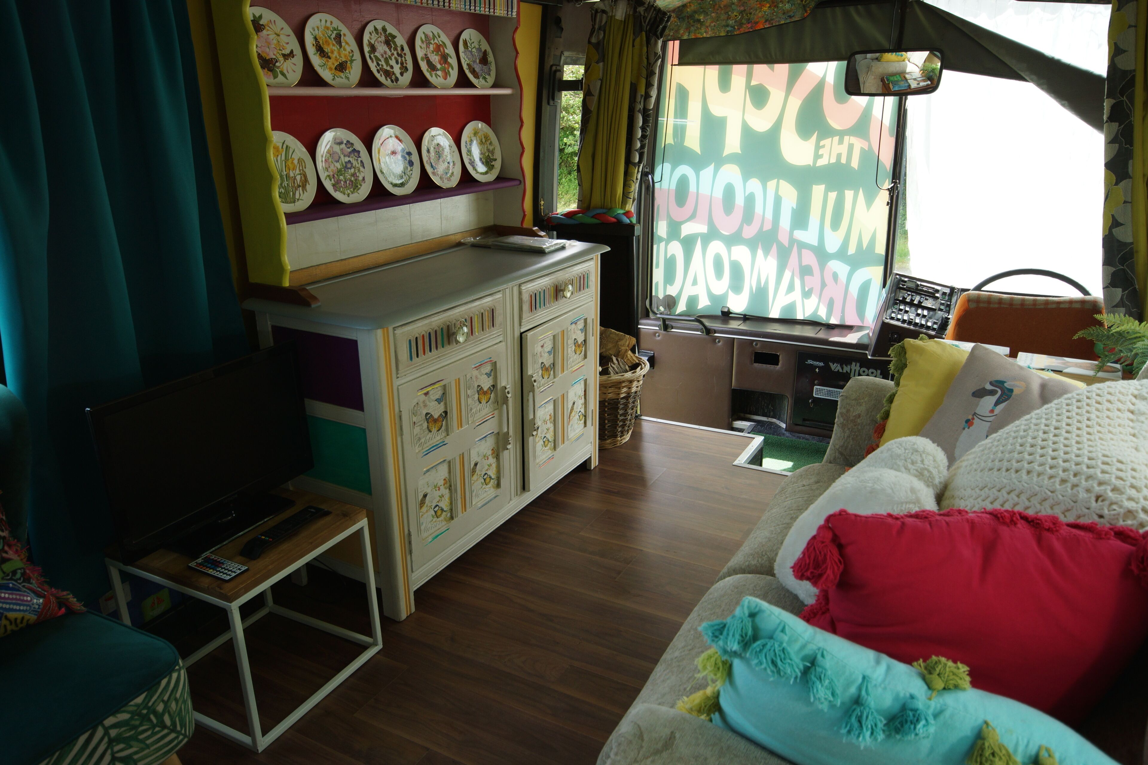 Un salon confortable avec un canapé, un téléviseur et un coin cuisine agrémenté d’une étagère où sont exposées des assiettes décoratives.