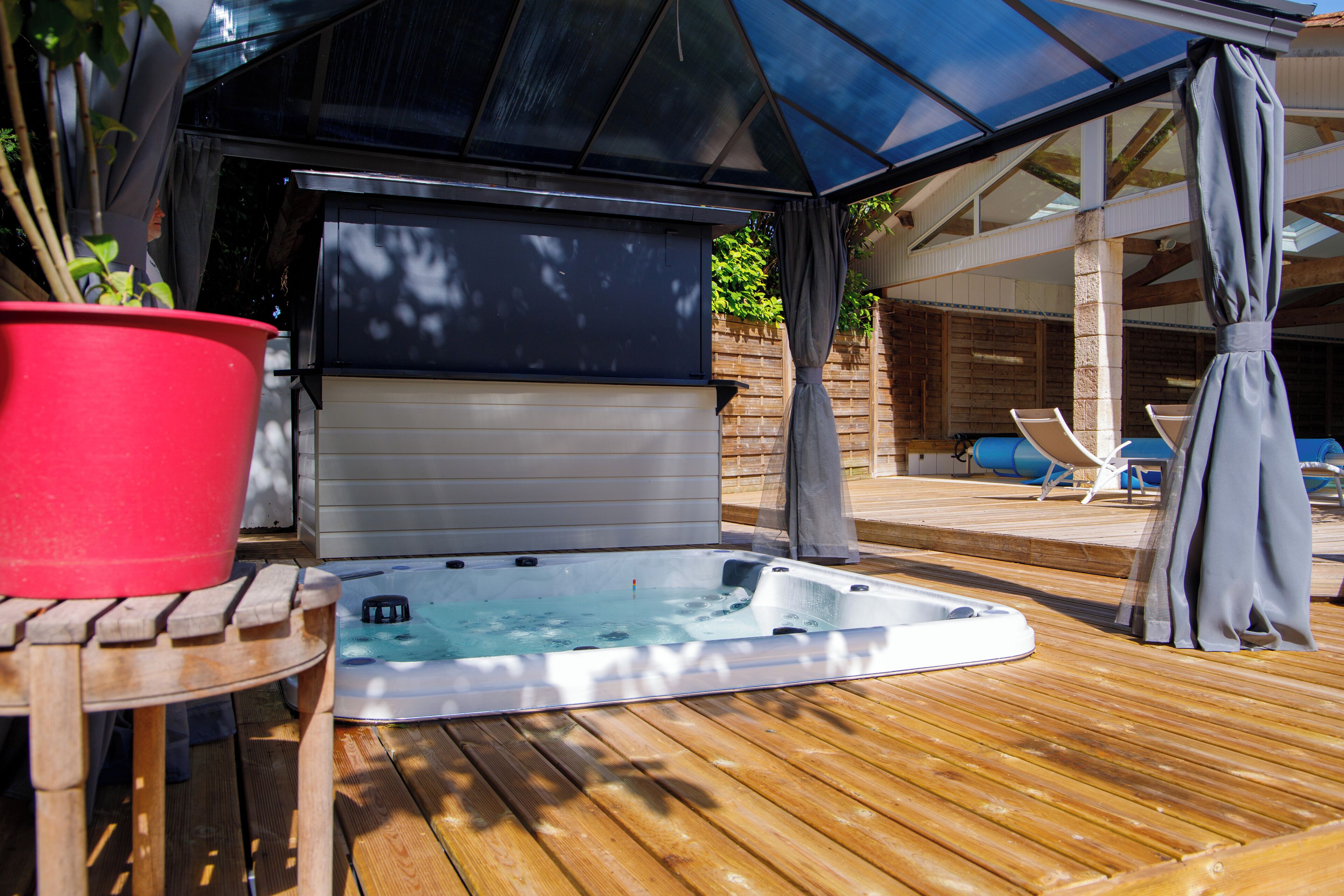 A wooden deck with a hot tub, a red pot, and a covered area with chairs.