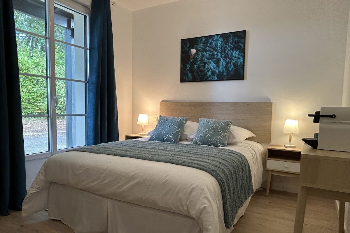 Ein ordentlich eingerichtetes Schlafzimmer mit einem großen Bett, zwei Nachttischen mit Lampen, einem Wandbild und einem Fenster mit Vorhängen.