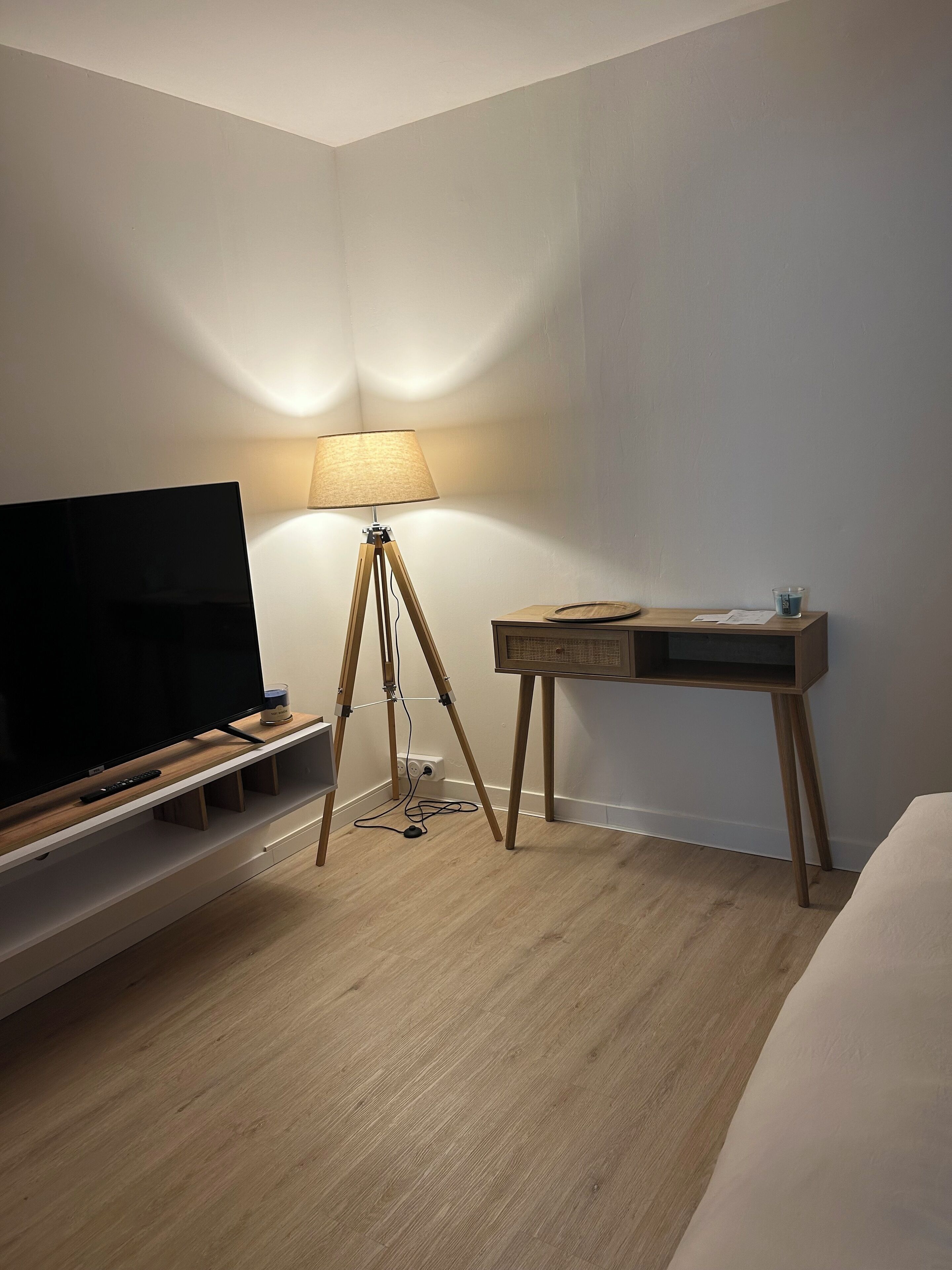 A hotel room with a wooden floor, a TV on a wooden stand, a desk with a lamp, and a bed.