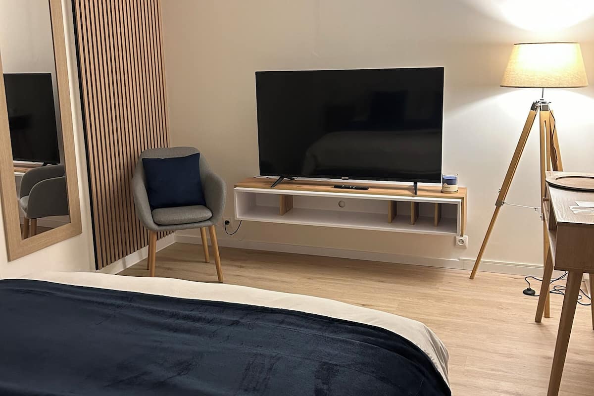 Ein modernes Hotelzimmer mit einem großen Bett, einem Fernseher auf einem Holzständer, einem Sessel, einer Lampe und einem Spiegel.