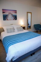 Cama bem arrumada com manta azul, cabeceira de madeira e duas almofadas estampadas. Há uma mesa de cabeceira com uma luminária e um espelho na parede. Uma poltrona azul-esverdeada é visível no canto.