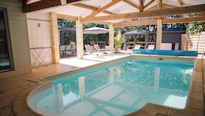 A covered swimming pool with lounge chairs and umbrellas, surrounded by a wooden deck and a brick wall.