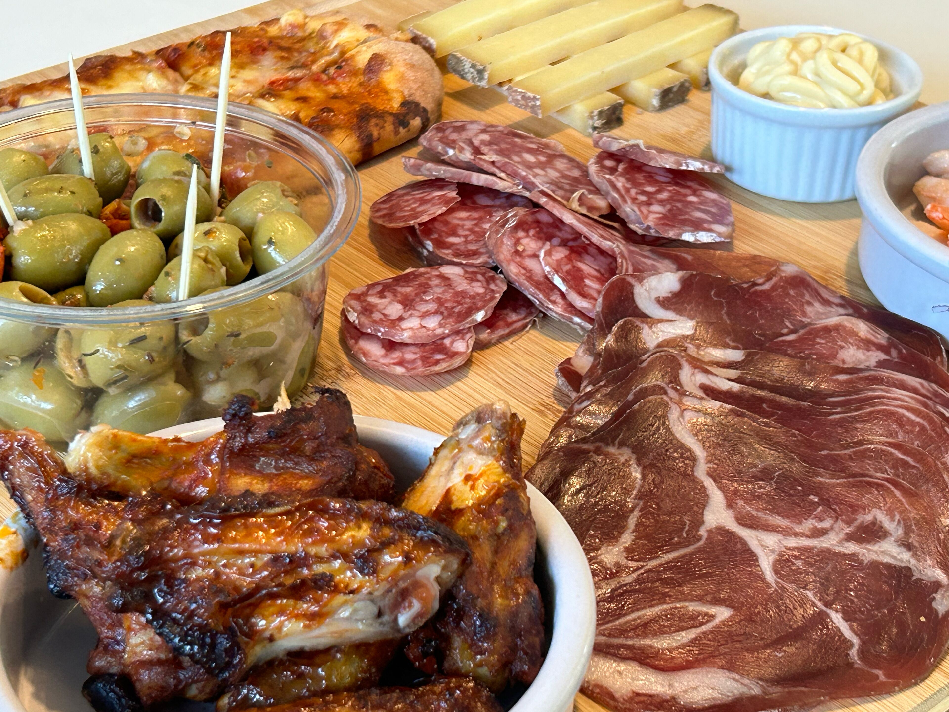 Diversos tipos de carne como salami en rodajas, embutidos y pollo a la parrilla, junto con aceitunas y queso sobre una tabla de madera.