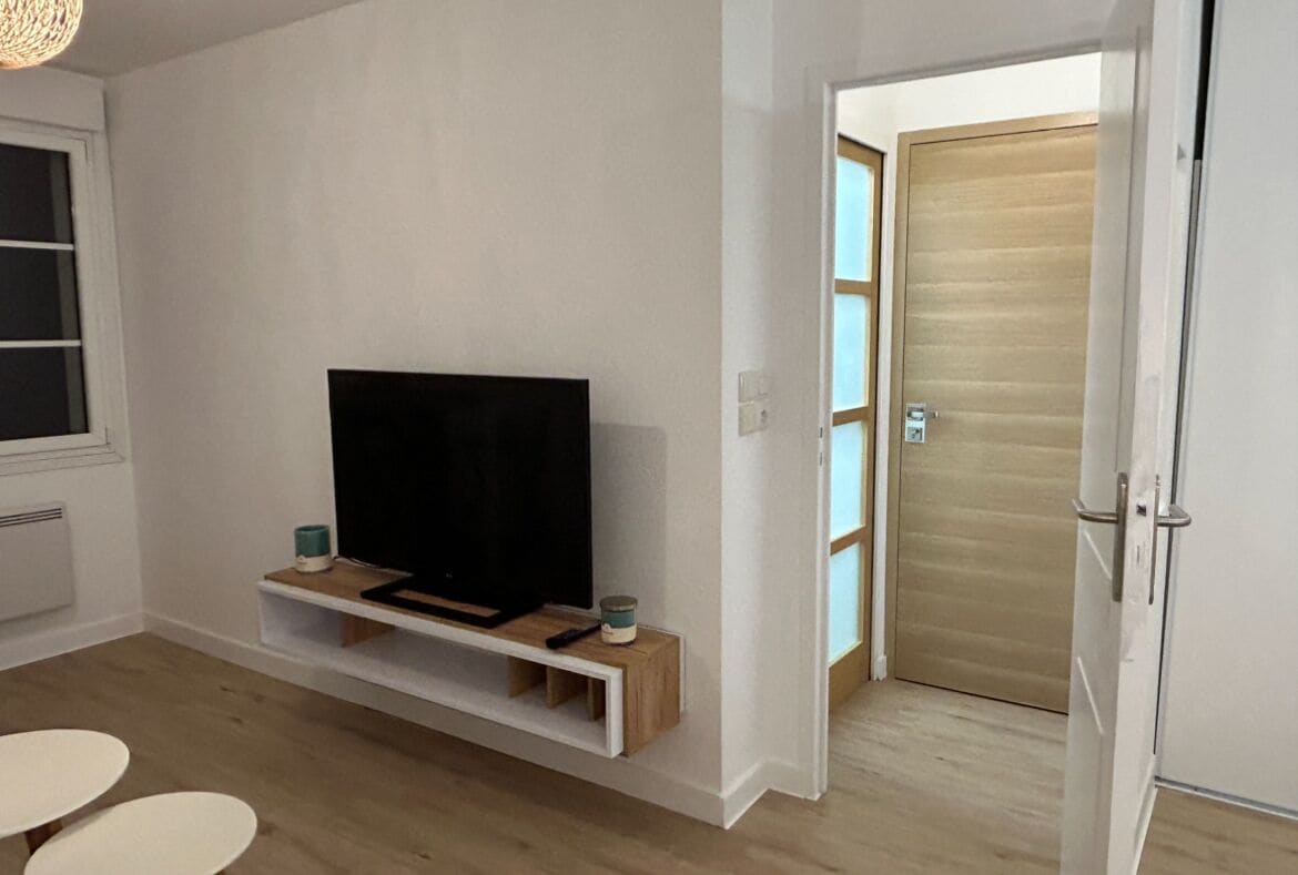 A modern living room with a flat-screen TV on a wooden shelf, a small table, and a door leading to another room.