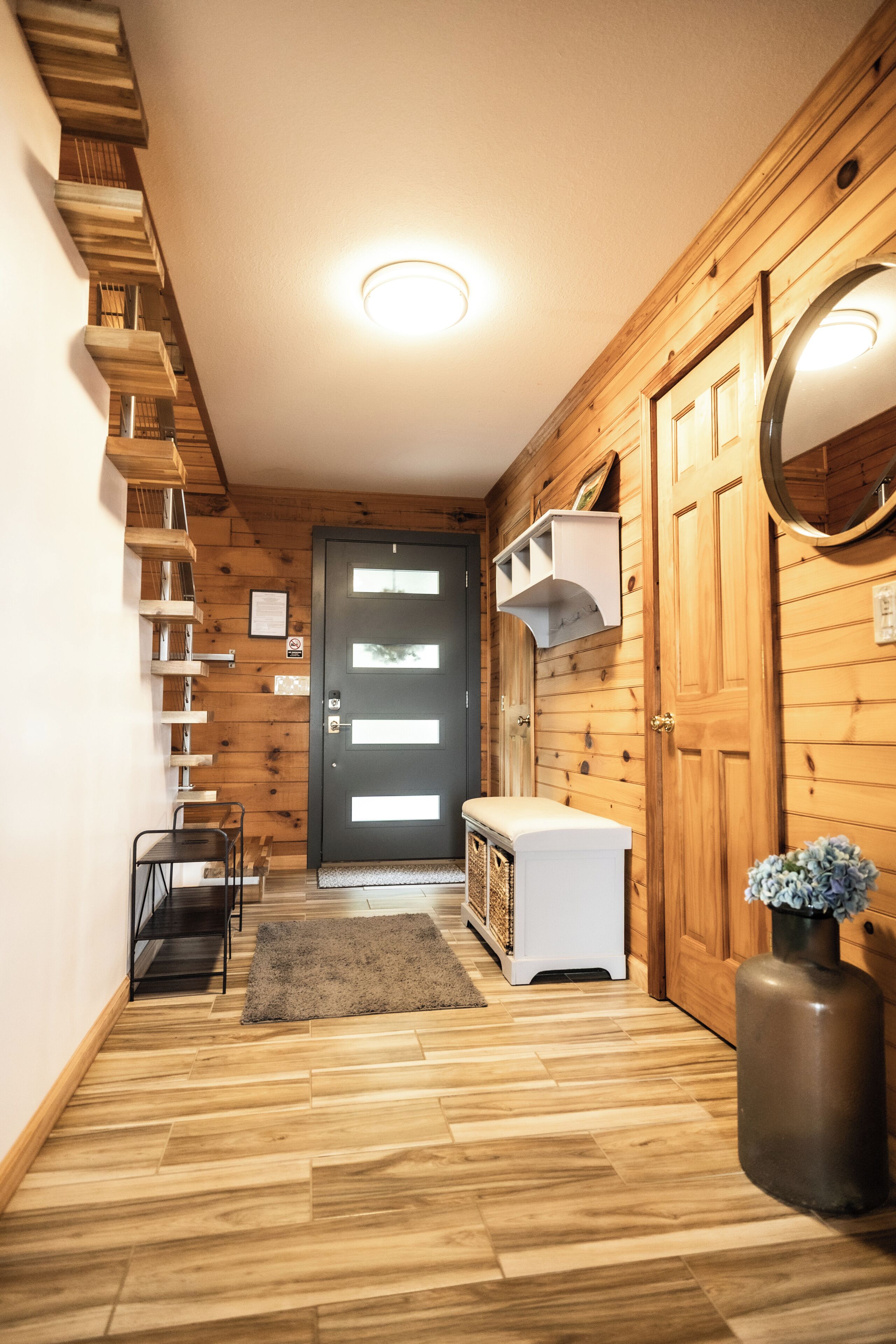 Un couloir aux murs en bois, une porte noire avec des panneaux de verre dépoli, un banc blanc, un miroir rond et une petite table avec des chaises.