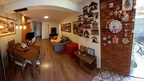 Een ruime woonkamer met een houten eettafel, een bank en een rode poef. Er hangen meerdere decoraties aan de muur, waaronder klokken en ornamenten in de vorm van huizen.