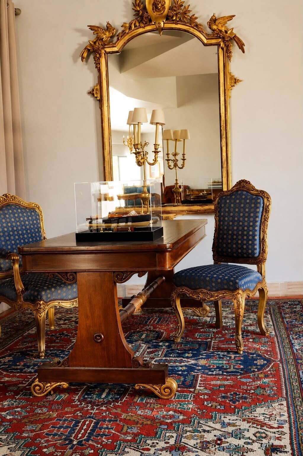 A room with a large ornate mirror, a wooden table, two blue upholstered chairs, and a patterned carpet.