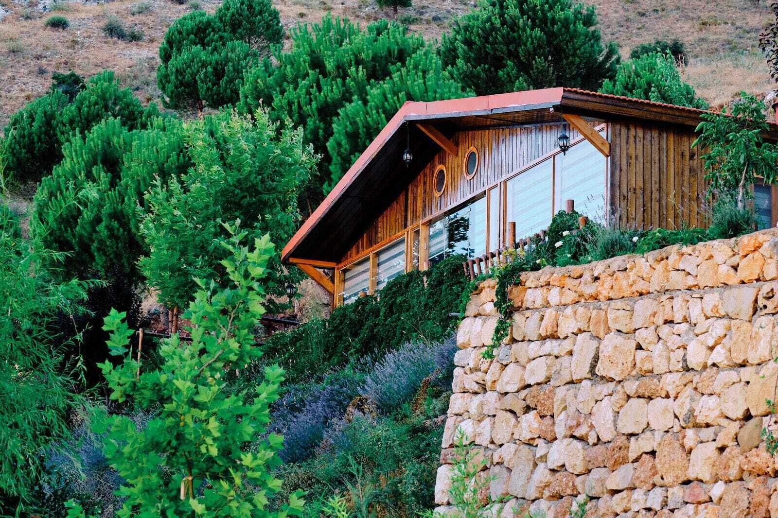 Une cabane en bois rustique, dotée d’un toit en pente, entourée d’une végétation luxuriante et d’un mur de pierres.