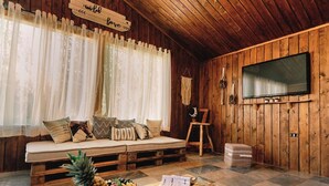 L’intérieur d’un chalet accueillant, avec un plafond en bois, un canapé agrémenté de coussins, un téléviseur et une table sur laquelle sont disposés des fruits et un carnet.