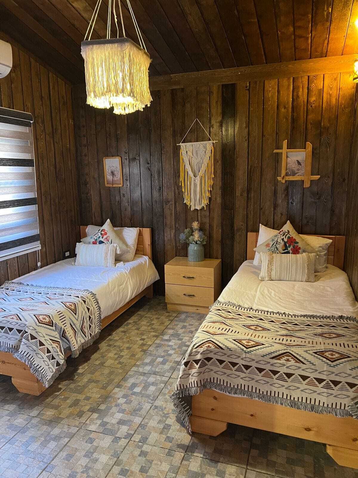 Ett rustikt trerum med två sängar, en hängande lampa i makramé och ett litet träbord vid sängen med en vas.