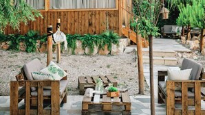 Un espace extérieur aménagé dans un style rustique, avec des meubles en bois et un sol carrelé en pierre, entouré de verdure et bordé d’un bâtiment en bois doté d’un toit rouge.