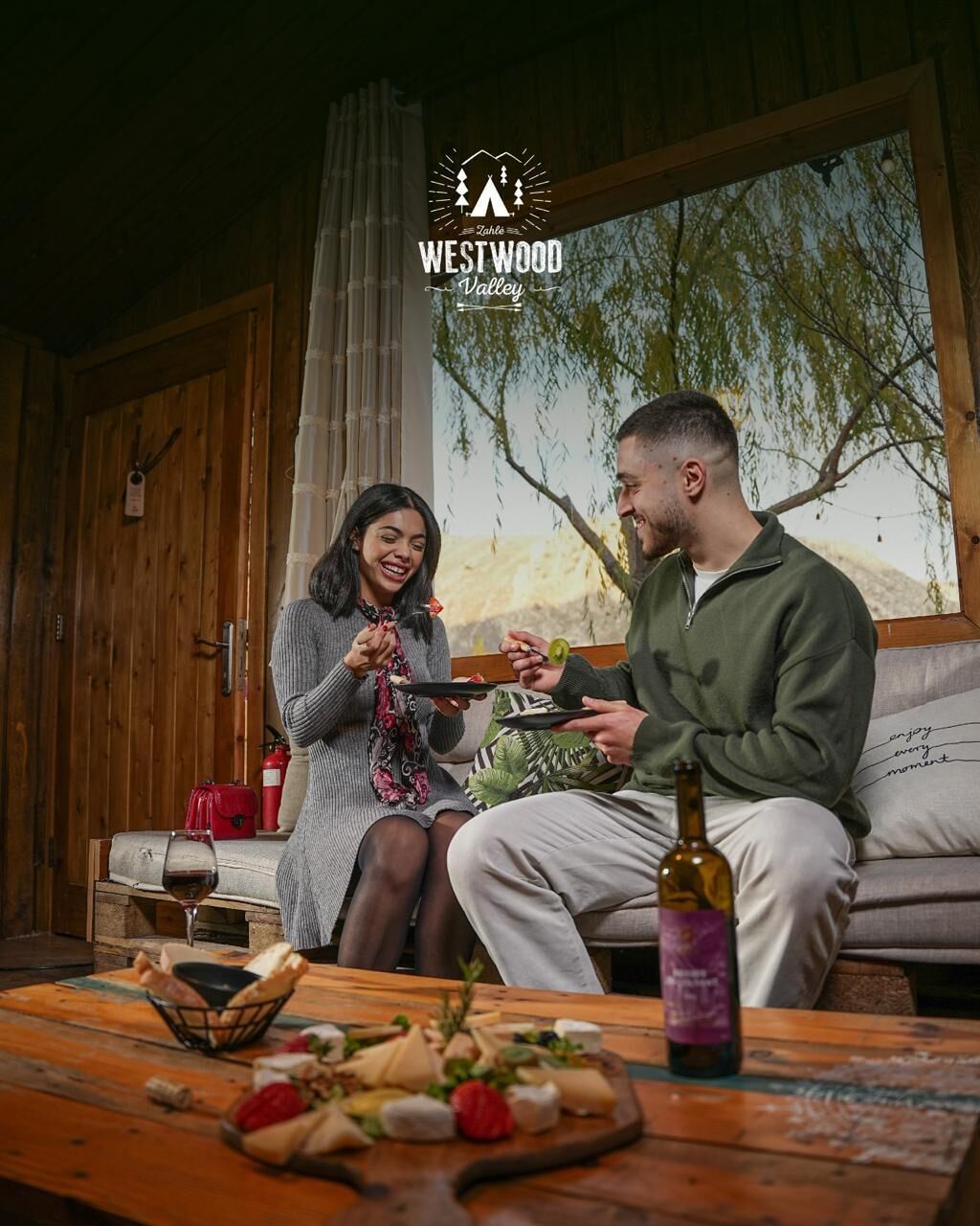 A couple sitting on a wooden bench in a rustic cabin with a table in front of them holding food and a bottle of wine.