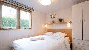 Ett sovrum med en säng, ett fönster, ett sängbord, en liten avlastningsyta, en garderob och en hylla med en vas och en hjärtformad prydnad.