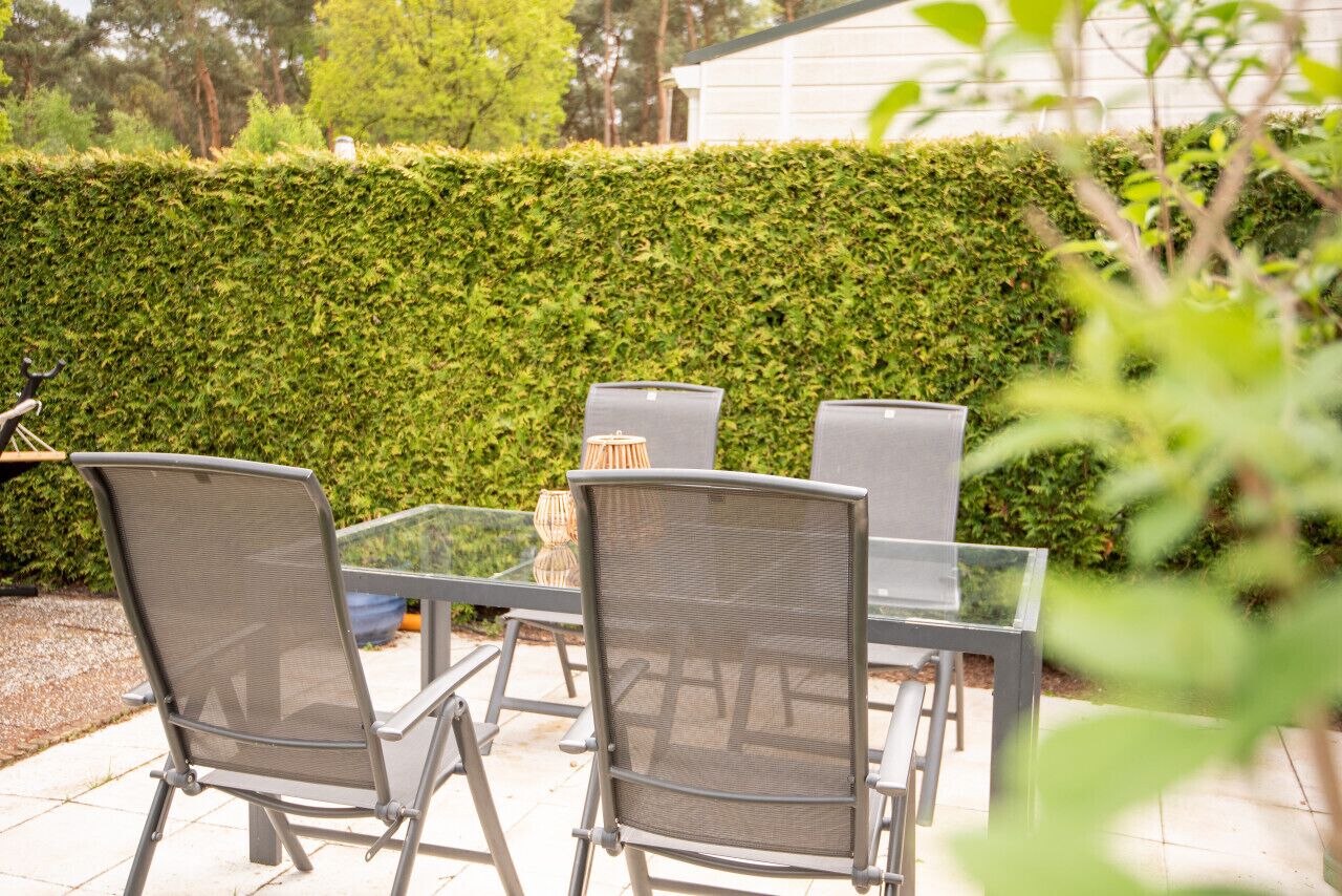 Une terrasse extérieure avec une table en verre et des chaises en métal, entourée d’un arbuste taillé avec soin.