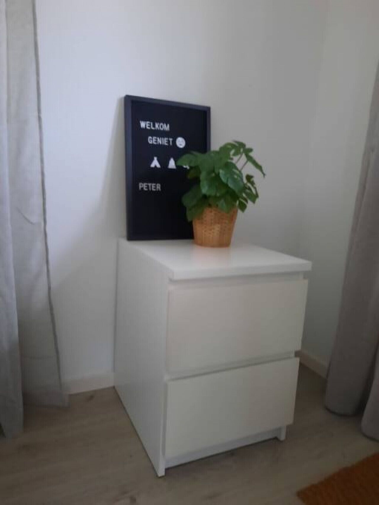 Una taula de nit blanca amb una planta en test i un cartell de benvinguda.