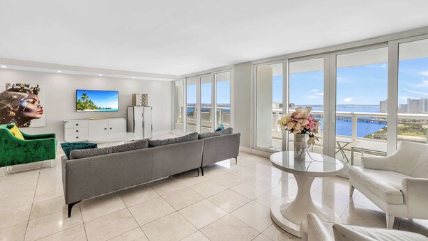 A modern living room with a grey sectional sofa, a white armchair, a round coffee table, and a television on a white cabinet. Large windows offer a view of a body of water and buildings. - 39th Floor Apt | Hot Tub | Gym | StayCozy (Miami)